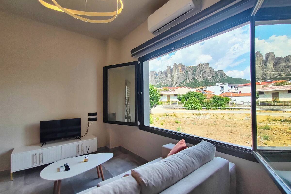 Een moderne kamer met een groot raam, een flatscreen-televisie, een witte bank en een kleine tafel.