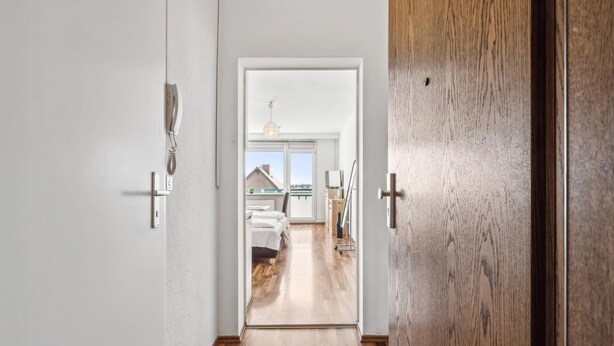 Een gang die leidt naar een slaapkamer met een bed, een bureau en een stoel. Er is een raam met uitzicht.