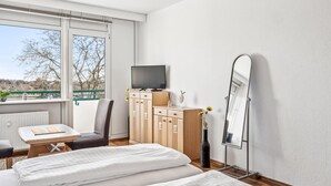 Een slaapkamer met een bed, een televisie, een dressoir, een spiegel en een raam met uitzicht op bomen en een groene balustrade.