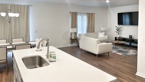 Eine moderne Küche mit Kochinsel, ein Essbereich mit Stühlen, ein Wohnzimmer mit Sofa und ein an der Wand montierter Fernseher.