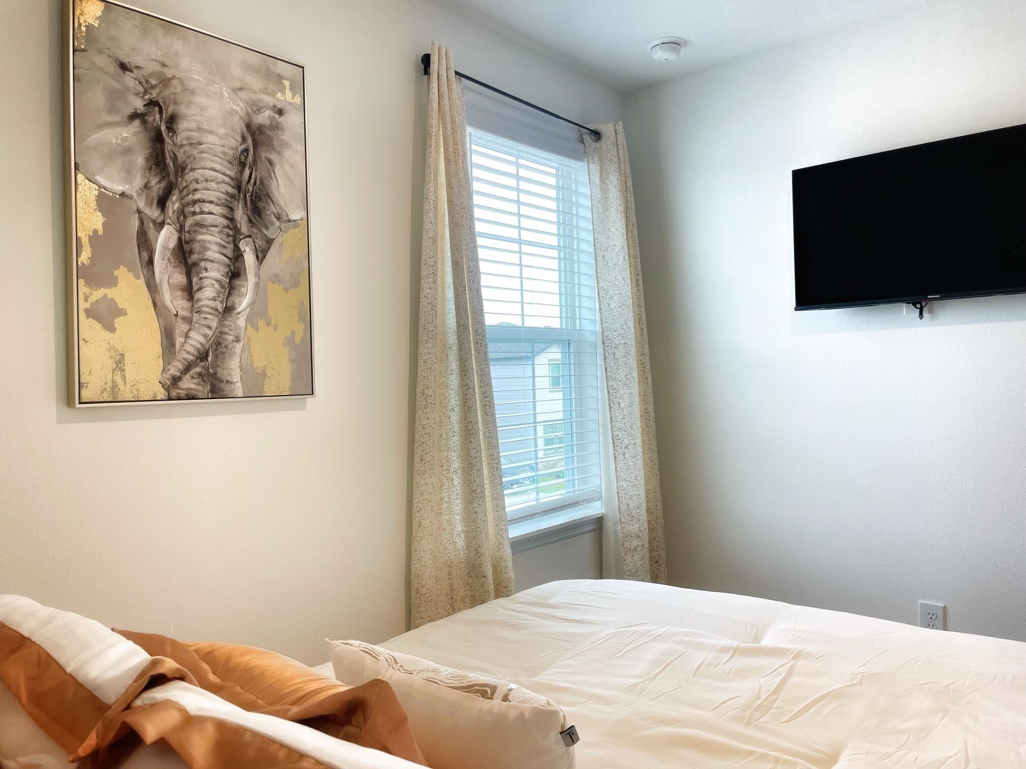 A bedroom with a bed, a painting of an elephant, a window with curtains, and a mounted TV.