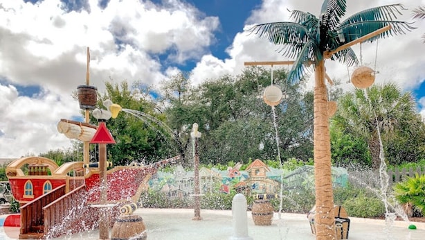 Ein Wasserpark mit einem thematisch gestalteten Spielbereich im Schiffsstil, einer Palme und einer Fontäne.