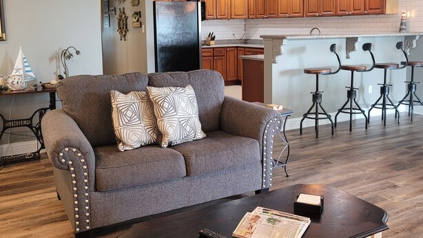 Una sala de estar con sofá y mesa de centro, una cocina con armarios de madera y refrigerador, y una zona de bar con taburetes.
