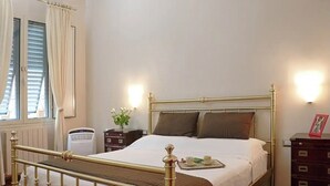 Una cama bien tendida con estructura metálica, una mesita de noche con lámpara y una ventana con cortinas.