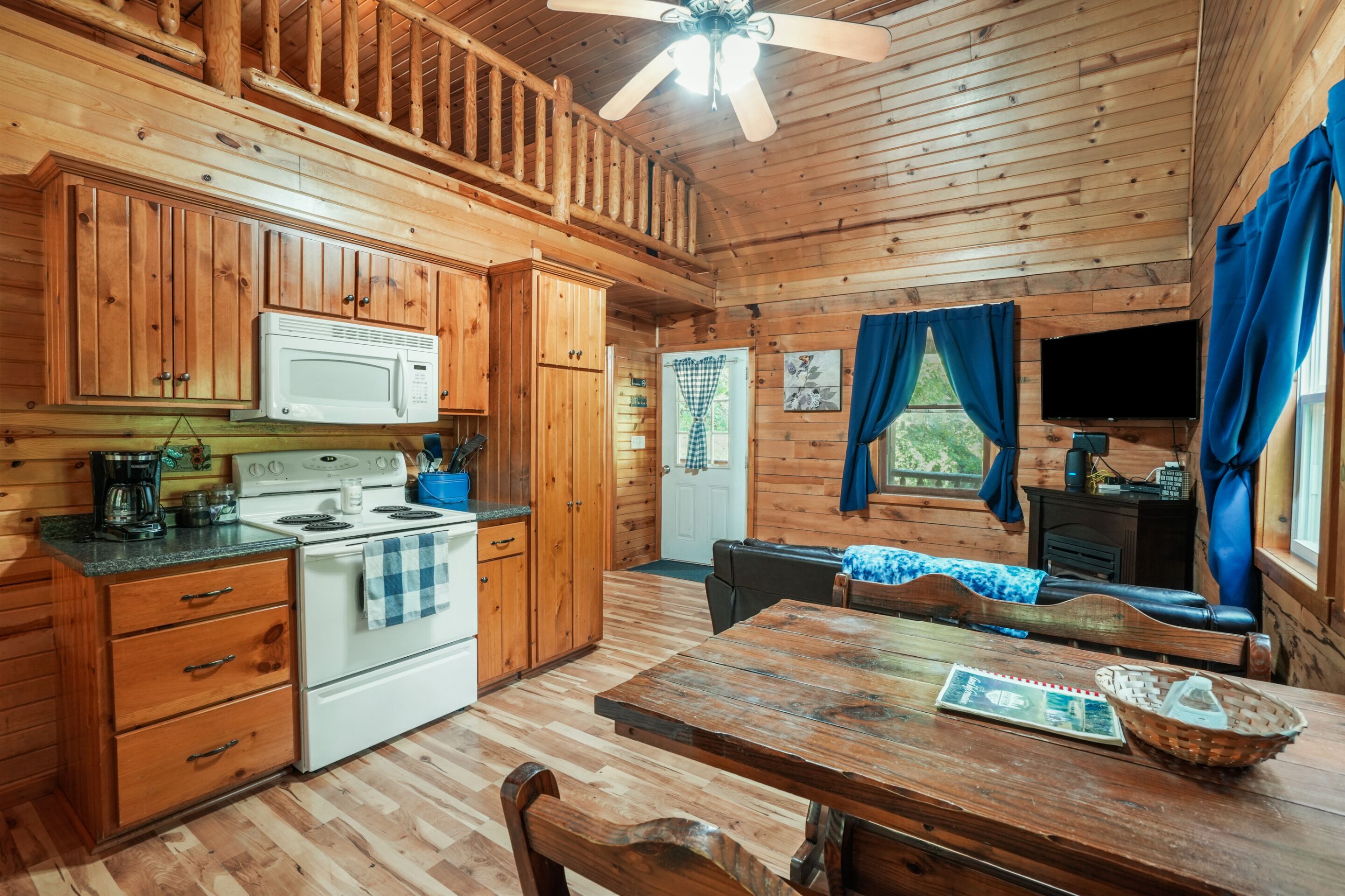 A wooden cabin with a kitchen featuring a stove, microwave, and cabinets. There is a dining table with chairs and a television in the corner.