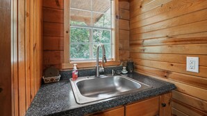 Une cuisine rustique aménagée dans une cabane en bois, avec un plan de travail en granit, un évier en acier inoxydable et une fenêtre donnant sur la forêt.