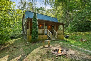 A rustic cabin with a porch, a fire pit, and a grill in a wooded area. - Butterfly Crossing | Private Lake, Hot Tub (Logan)