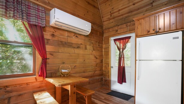 A wooden cabin interior with a wooden table, bench, white refrigerator, and an air conditioning unit. - Fireflies Hollow | Romantic Pond Retreat, Hot Tub (McArthur)