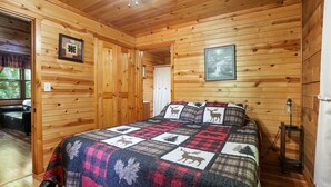 A wooden cabin bedroom with a bed, a framed picture on the wall, and a window with a view of trees. - Fireflies Hollow | Romantic Pond Retreat, Hot Tub (McArthur)