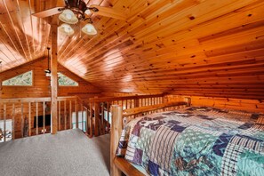 A wooden cabin interior with a bed, a ceiling fan, and a view of the outdoors.