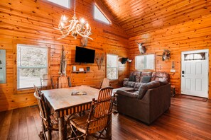 A log cabin interior with a dining table, chairs, a sofa, a TV, and mounted animal heads.