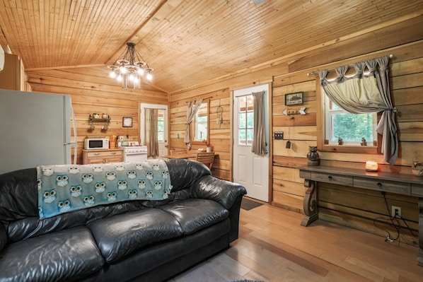 A cozy cabin interior with a wooden ceiling, a black leather sofa, a wooden dining table, and a kitchen area.