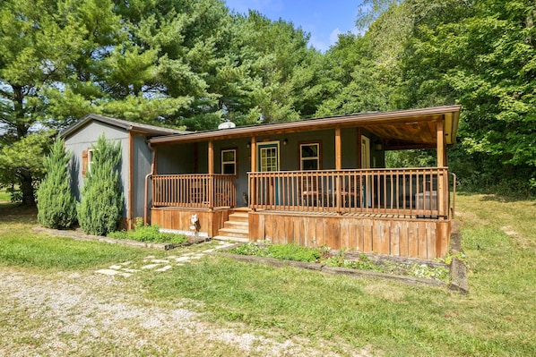 A two-story wooden cabin with a porch, surrounded by trees and a grassy yard.