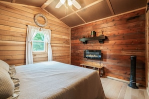 A wooden cabin bedroom with a bed, a fireplace, a ceiling fan, and a window with curtains.