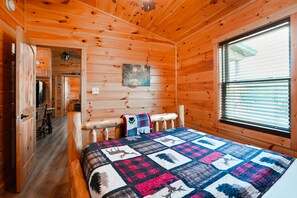 A wooden cabin interior with a bed, a window, and a door leading to a kitchen.