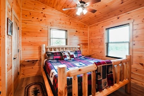 A wooden cabin bedroom with a bed, a ceiling fan, and two windows.