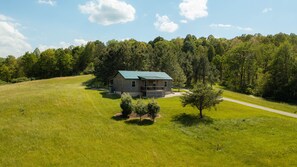 Une cabane en bois avec un toit vert, entourée d’un champ herbeux et d’une colline boisée.