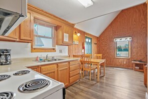 A kitchen with wooden cabinets, a stove, a sink, and a dining area with chairs. - Retreat Cabin #1 | Hocking Hills, Sleeps 6 (Logan)