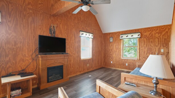 Ein gemütliches Zimmer mit Holzwänden, einem Deckenventilator, einem Kamin, einem Fernseher und einem Fenster mit Blick auf Grünflächen.