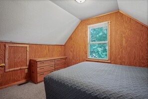 A bedroom with a sloped ceiling, wooden walls, a window with blinds, a bed with a quilted bedspread, and a wooden dresser.