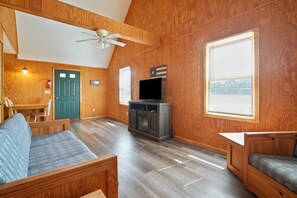 A cozy living room with a wooden floor, a ceiling fan, a television, and a green door.