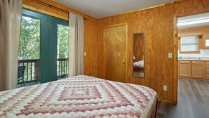A bedroom with a large bed, wooden walls, a sliding glass door to a balcony, and a small bathroom with wooden cabinets. - Retreat Cabin #2 | Hocking Hills,  Pet Friendly (Logan)