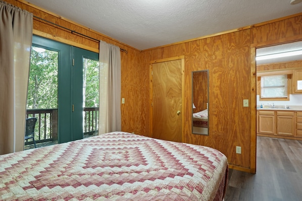 Un dormitorio con una cama grande, paredes de madera, un ventanal corredero a un balcón y un baño pequeño con armarios de madera.