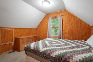 A bedroom with a wooden sloped ceiling, a bed with a patterned quilt, a wooden dresser, and a window with curtains.