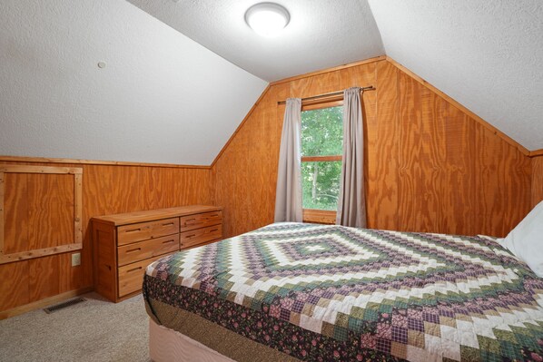 A bedroom with a wooden sloped ceiling, a bed with a patterned quilt, a wooden dresser, and a window with curtains.