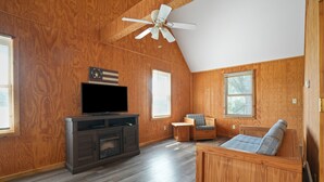 A spacious living room with wooden walls and flooring, a ceiling fan, a flat-screen TV, a wooden bench with cushions, and two windows with blinds. - Retreat Cabin #2 | Hocking Hills,  Pet Friendly (Logan)