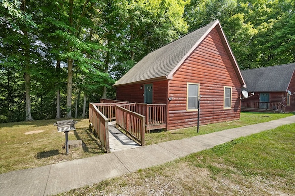 A red log cabin with a wooden deck and a small porch, surrounded by trees. - Retreat Cabin #8 | Hocking Hills, Fire Ring (Logan)