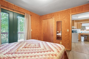 A bedroom with a large bed, wooden walls, a sliding door to a balcony, and a view of a kitchen. - Retreat Cabin #8 | Hocking Hills, Fire Ring (Logan)