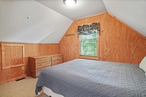 Une chambre avec un plafond en pente, des murs en bois, une fenêtre avec des stores, un lit recouvert d’une couverture matelassée et une commode en bois.