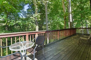 A wooden deck with a glass table and two chairs, surrounded by tall trees.
