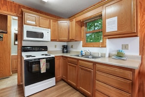 A kitchen with wooden cabinets, a white stove, a microwave, a sink, and a window with a view of trees.