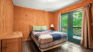 Een slaapkamer met een houten vloer, een bed met een gebloemd deken, een nachtkastje, een lamp en een schuifdeur naar een balkon.