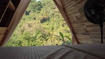 Habitación de madera triangular con cama y ventilador, con vistas a una exuberante ladera verde.