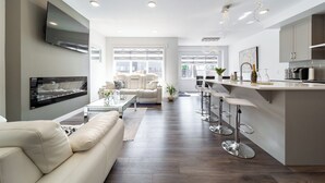 Una sala moderna con sofá, una mesa de centro y un televisor montado en la pared. Hay una zona de cocina con barra y taburetes.