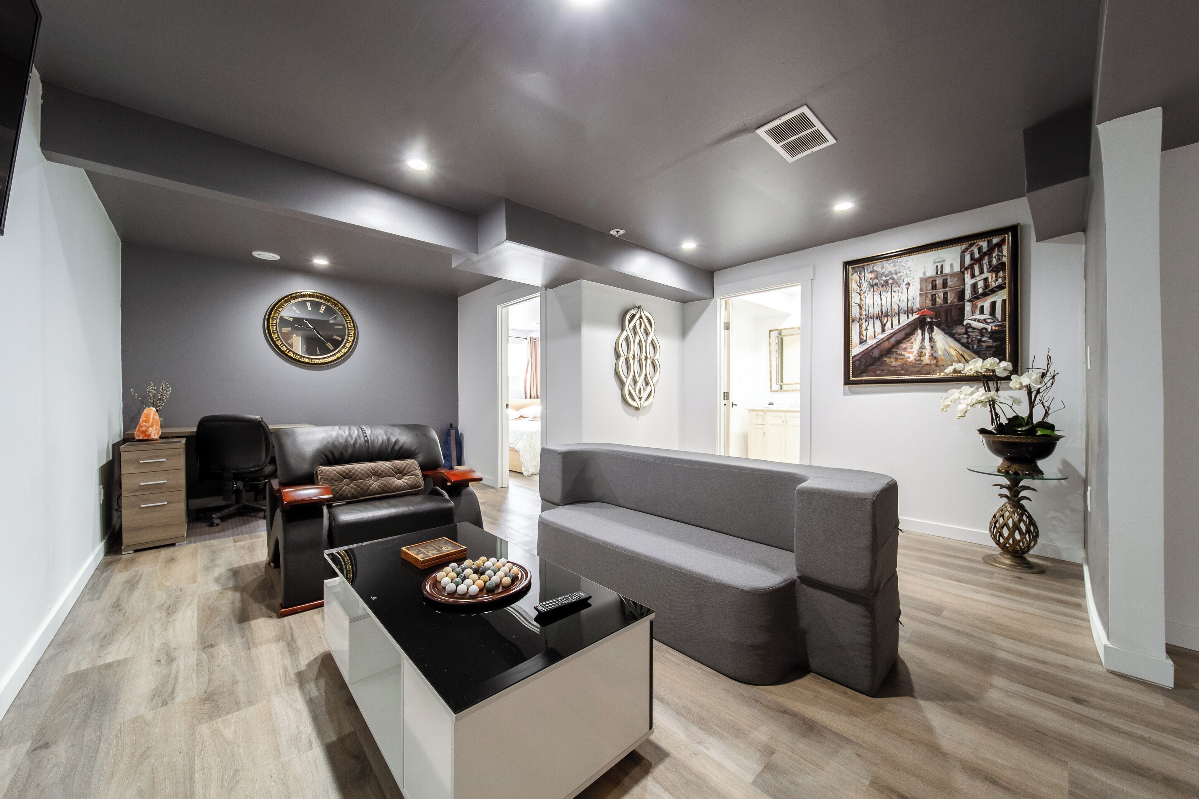 A modern living room with a grey sofa, a black coffee table, and a black leather chair. There is a painting on the wall and a clock on the ceiling.