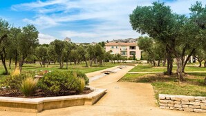 Un chemin menant à un bâtiment doté d’un toit en tuiles, entouré d’arbres et d’un jardin aménagé.