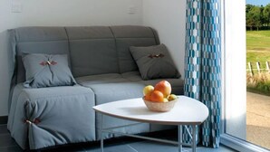 Une pièce moderne avec un canapé gris, une table ronde sur laquelle est posé un panier de fruits, et une vue sur un paysage verdoyant, visible à travers une grande fenêtre ornée de rideaux à motifs.