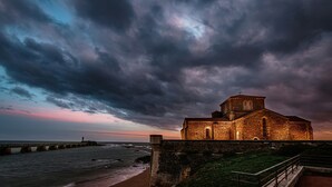 Una iglesia costera de fachada de piedra, un faro distante y un cielo tormentoso.