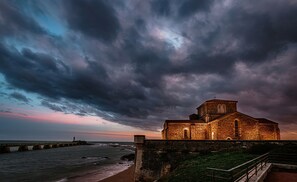 A coastal church with a stone facade, a lighthouse in the distance, and a stormy sky.
