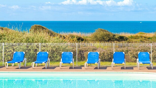 Zona de piscina con seis sillas reclinables azules alineadas, una valla blanca y vista al mar.