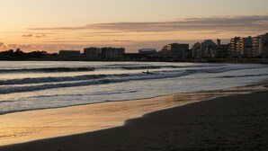 Una playa con olas, una persona practicando surf y edificios al fondo.