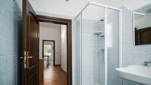 Een badkamer met een douche, een wastafel en een deur naar een andere kamer.