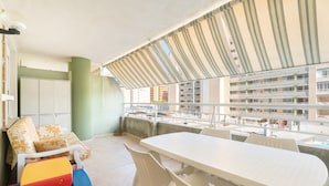 Een balkon met een witte tafel en stoelen, een bank met gekleurde kussens en uitzicht op een meerhoog gebouw.