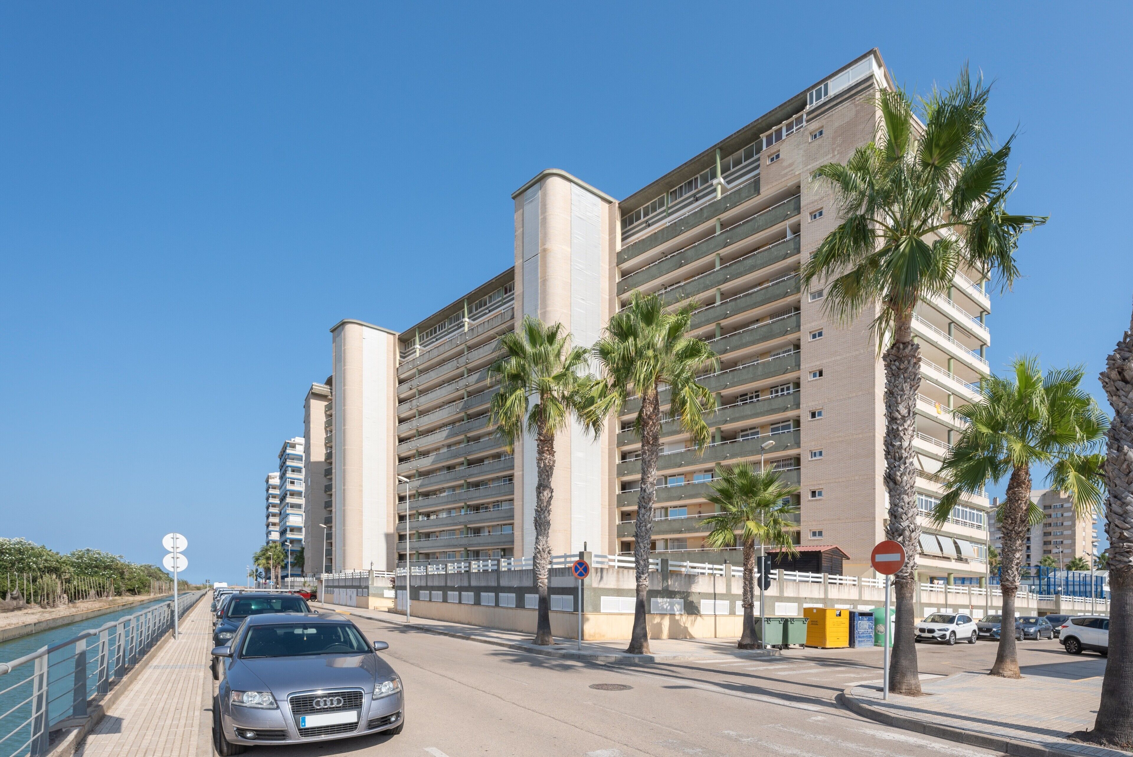 Un edificio de varios pisos con balcones, una pasarela peatonal y automóviles estacionados.