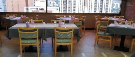 A dining area with tables and chairs, large windows with a city view, and a tiled floor.