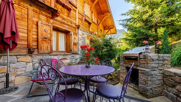 A wooden chalet with a stone fireplace, outdoor seating, and a view of the surrounding greenery.